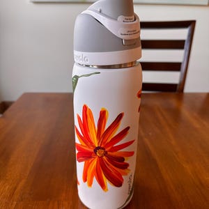 Puede incluir: Botella de agua aislada blanca con tapa gris y un vibrante dise&ntilde;o floral naranja y amarillo. La botella tiene un bot&oacute;n y una etiqueta que dice "Lift to unlock". Decorada con un colibr&iacute; y la marca "owala".