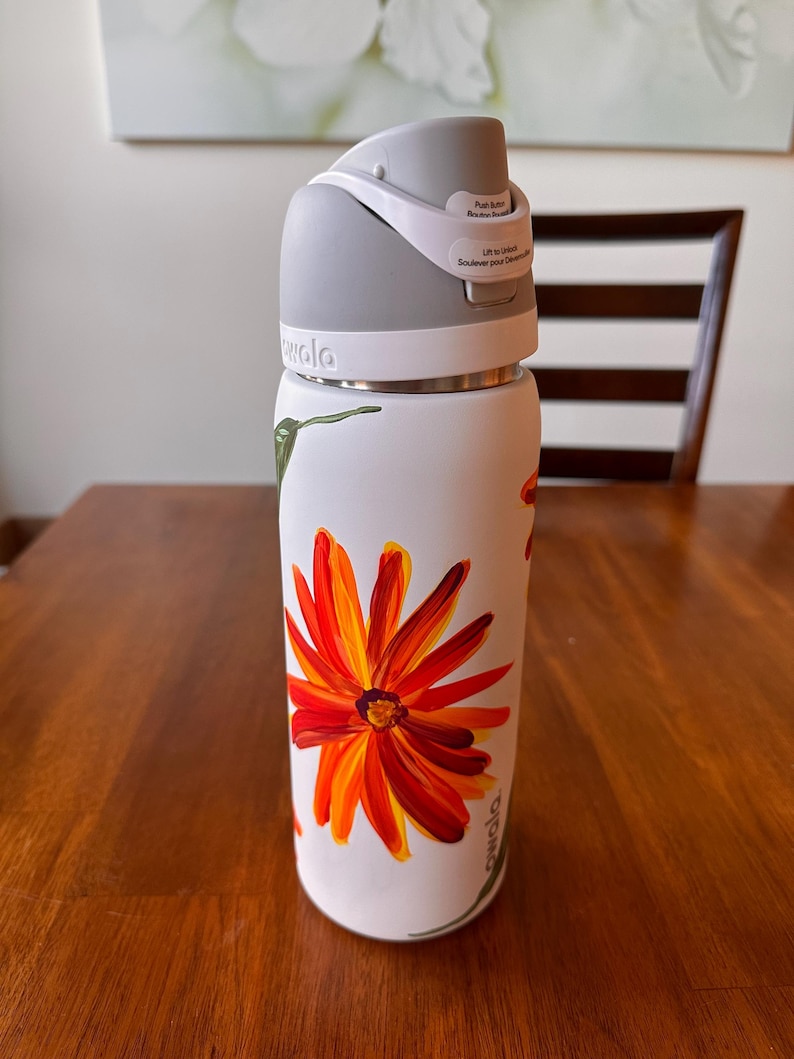 Puede incluir: Botella de agua aislada blanca con tapa gris y un vibrante dise&ntilde;o floral naranja y amarillo. La botella tiene un bot&oacute;n y una etiqueta que dice "Lift to unlock". Decorada con un colibr&iacute; y la marca "owala".
