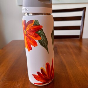 Puede incluir: Una botella de agua aislada blanca con asa y tapa grises. La botella est&aacute; decorada con flores naranjas y rojas pintadas a mano y hojas verdes. La botella est&aacute; sobre una superficie de madera.