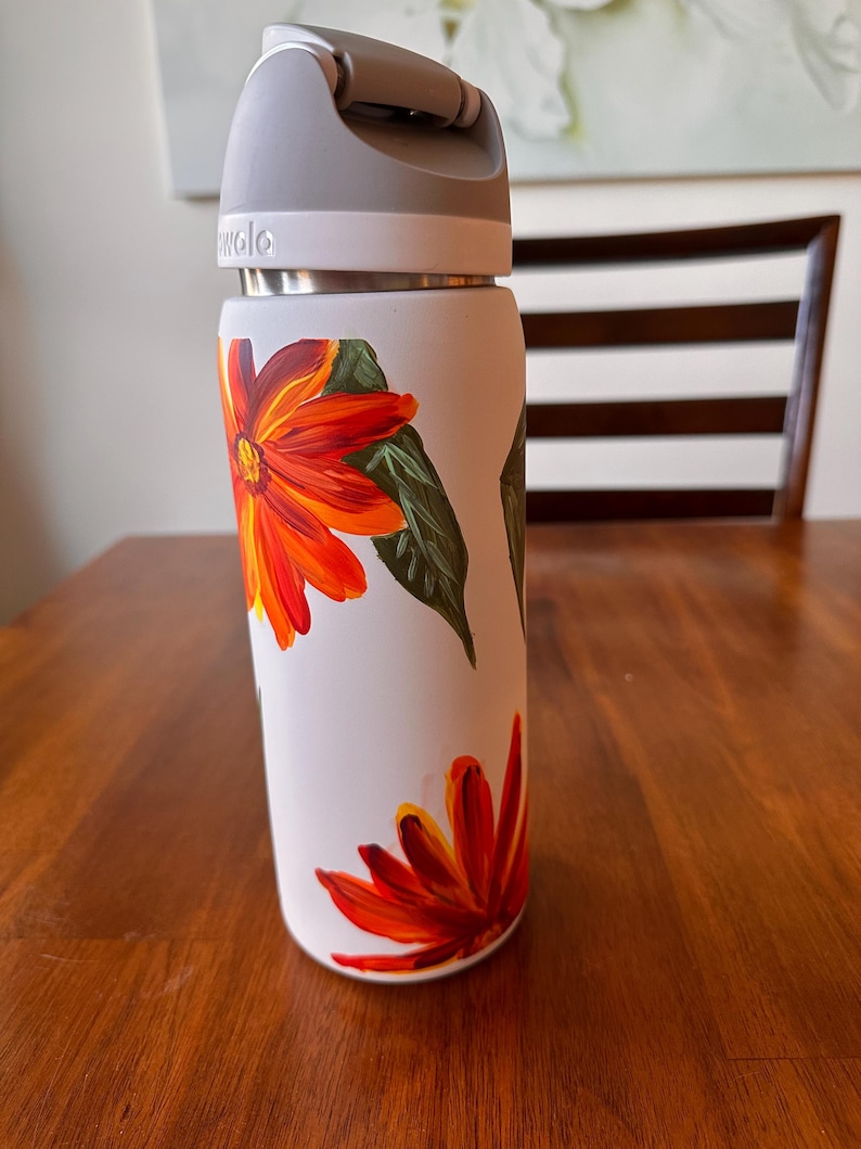 Puede incluir: Una botella de agua aislada blanca con asa y tapa grises. La botella est&aacute; decorada con flores naranjas y rojas pintadas a mano y hojas verdes. La botella est&aacute; sobre una superficie de madera.