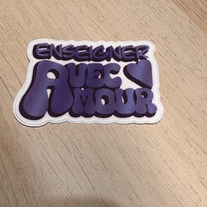 May include: A sticker with the words "ENSEIGNER AVEC AMOUR" in purple, with a white border. The text is in a stylized font with a heart symbol. The sticker is on a light brown wooden surface.