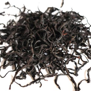 May include: A close-up of a pile of dark, dried tea leaves. The leaves are a deep reddish-brown colour, with some lighter brown and tan hues. The leaves have a twisted, wiry texture, and are scattered against a white background.