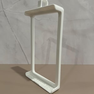 May include: A white, rectangular 3D-printed object with a handle on top and a rectangular frame. The base of the object has the text "COLLECT3D.COM" printed on it. The object is on a brown surface.