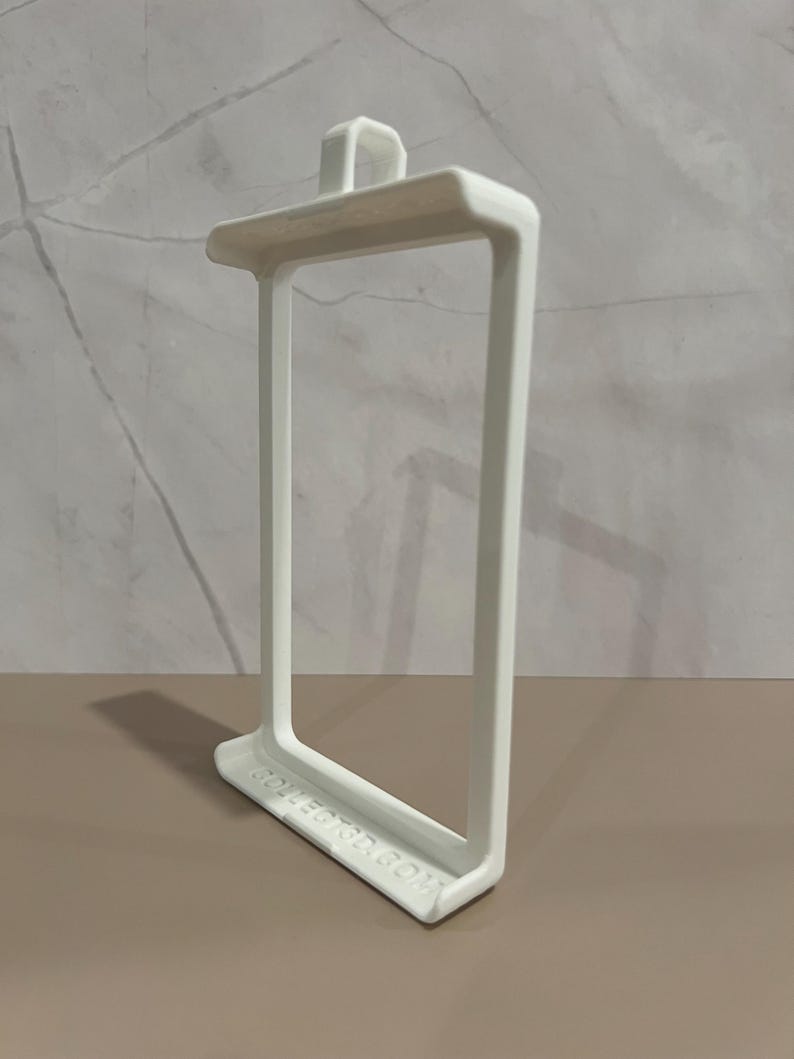 May include: A white, rectangular 3D-printed object with a handle on top and a rectangular frame. The base of the object has the text "COLLECT3D.COM" printed on it. The object is on a brown surface.
