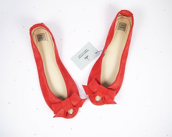 May include: A pair of red suede ballet flats with a bow detail on the toe. The flats are open-toed and have a simple design.