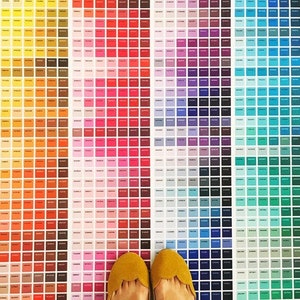 Puede incluir: Una gran carta de colores con cientos de pequeños cuadrados de diferentes colores, dispuestos en un patrón de arcoíris. La carta está en el suelo y un par de zapatos amarillos están de pie sobre ella.