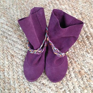 May include: A pair of purple suede ankle boots with colorful braided straps. The boots have a slouchy fit and a rounded toe.