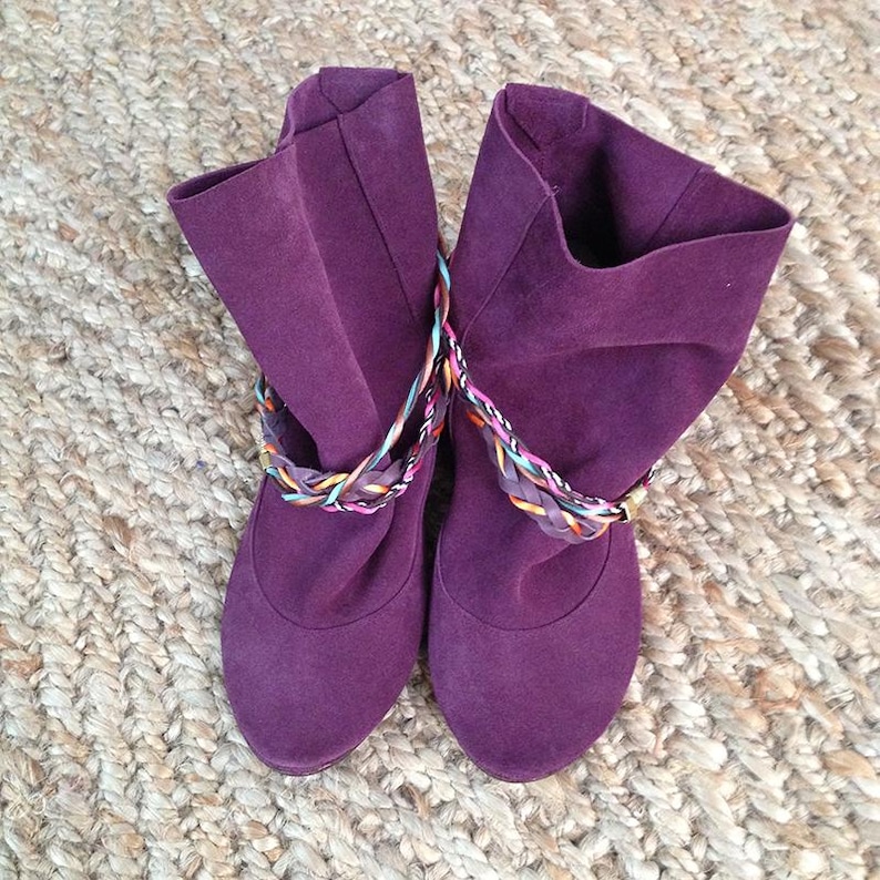 May include: A pair of purple suede ankle boots with colorful braided straps. The boots have a slouchy fit and a rounded toe.