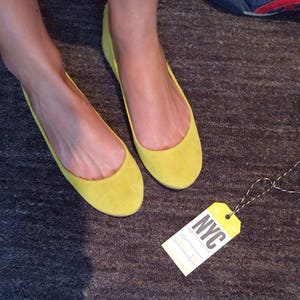 May include: A pair of bright yellow suede ballet flats with a simple design. The flats are on a grey carpet with a luggage tag that says "NYC" on it.
