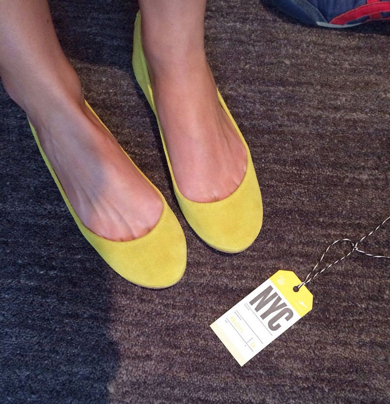 May include: A pair of bright yellow suede ballet flats with a simple design. The flats are on a grey carpet with a luggage tag that says "NYC" on it.