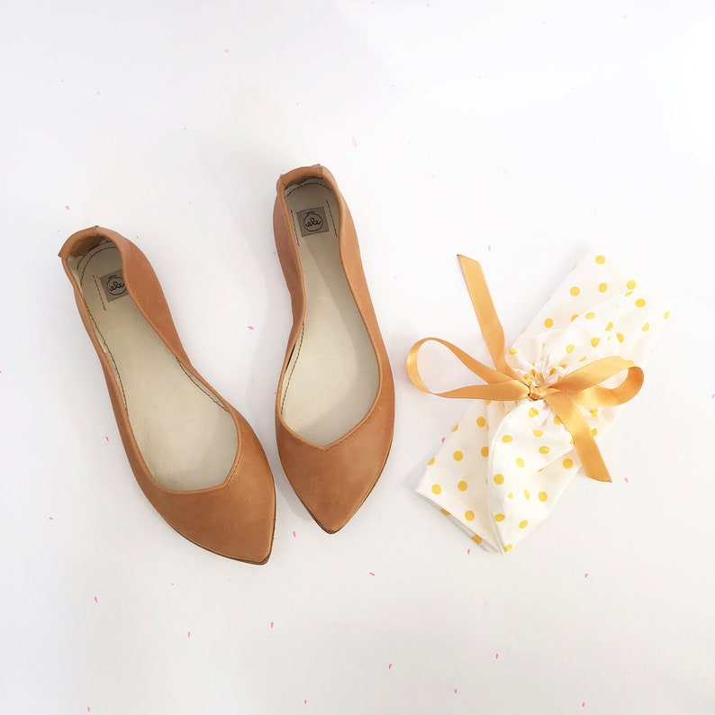 May include: Two pairs of brown leather ballet flats with pointed toes. The flats are on a white surface with a white and yellow polka dot fabric with a gold ribbon tied around it.