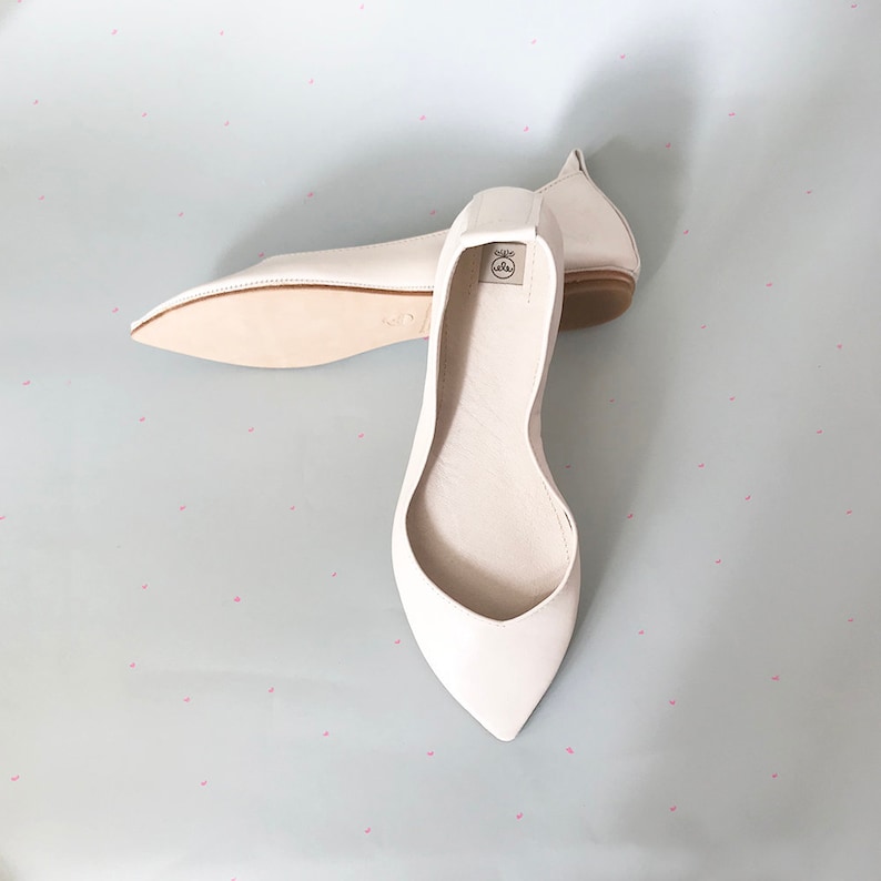 May include: A pair of white leather ballet flats with pointed toes. The flats are laid out on a light blue background with a white polka dot pattern.