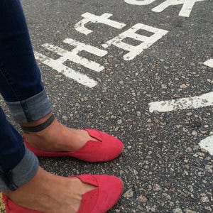 May include: A pair of red suede flats with a pointed toe and a small cut-out detail on the vamp. The shoes are worn by a person standing on a paved surface with the words "LOOK HERE" painted in white.