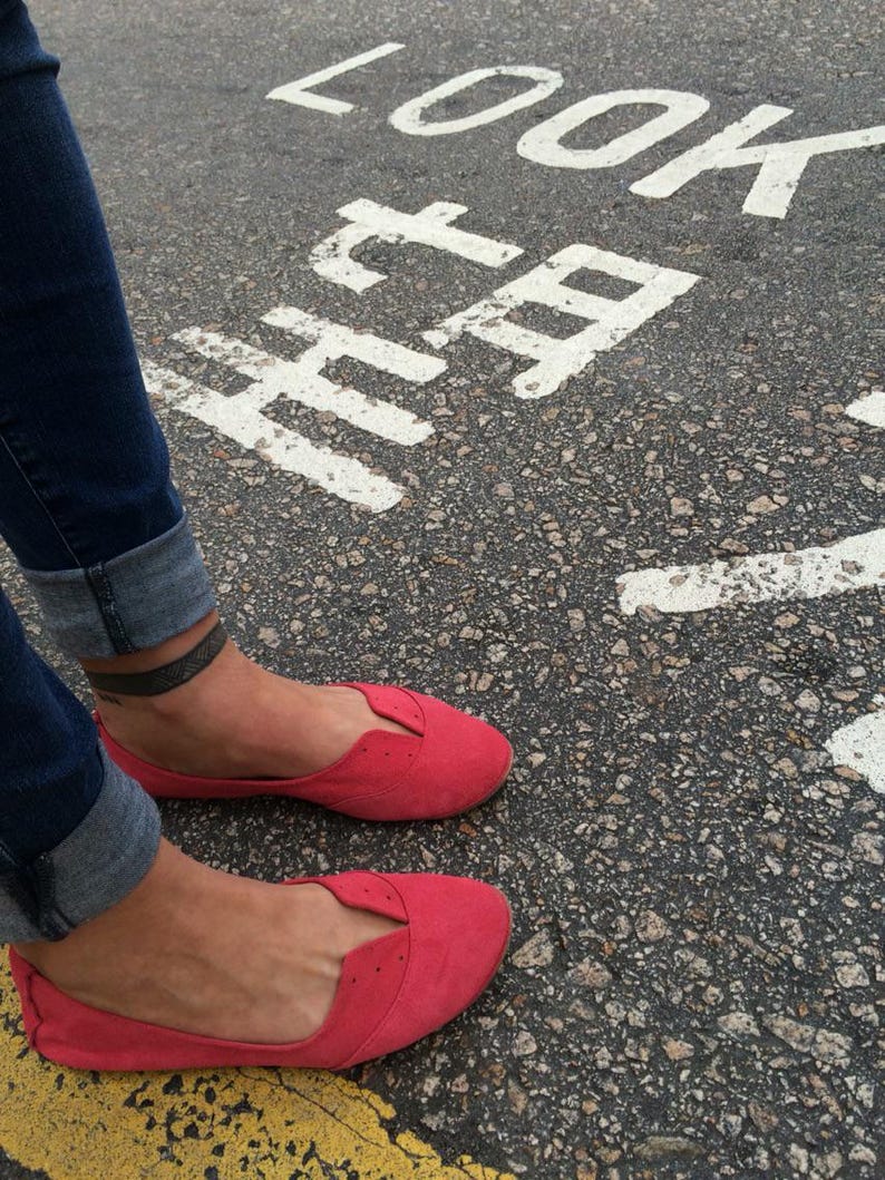 May include: A pair of red suede flats with a pointed toe and a small cut-out detail on the vamp. The shoes are worn by a person standing on a paved surface with the words "LOOK HERE" painted in white.