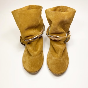 May include: A pair of brown suede ankle boots with a braided strap detail around the ankle.