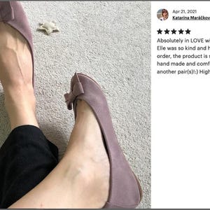 May include: A pair of mauve ballet flats with a bow on the toe. The shoes are made of suede and have a comfortable fit.