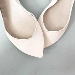 May include: A pair of white leather ballet flats with pointed toes. The flats are on a light blue background with pink hearts.