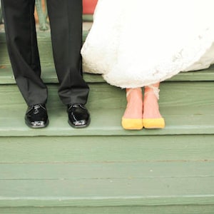 May include: A bride and groom stand on a set of green wooden steps. The bride is wearing a white wedding dress and yellow shoes. The groom is wearing a black suit and black shoes.