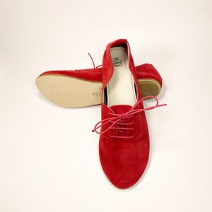 May include: A pair of red suede lace-up flats with a white leather lining. The shoes have a pointed toe and a low heel.