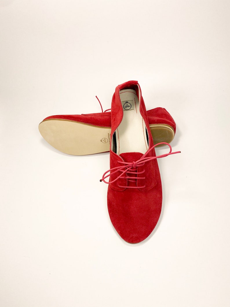 May include: A pair of red suede lace-up flats with a white leather lining. The shoes have a pointed toe and a low heel.