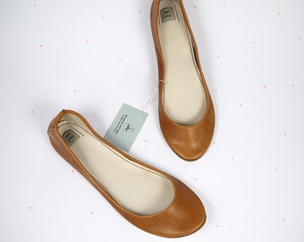 May include: A pair of tan leather ballet flats with a rounded toe and a light-coloured interior lining. Each shoe features a small interior brand label. A pale green rectangular tag with the text "Danskah-vintage leather accessories" is attached.