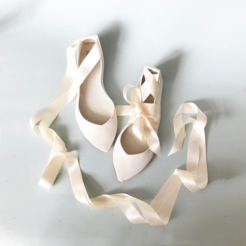 May include: A pair of ivory satin bridal flats with long ivory satin ribbons tied into a bow on one shoe. The shoes have a pointed toe.