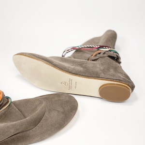 May include: A pair of gray suede ankle boots with colorful laces. The boots have a flat sole and a rounded toe. The text on the sole reads "Handmade and Design Luxury Accessories VFB 2020".