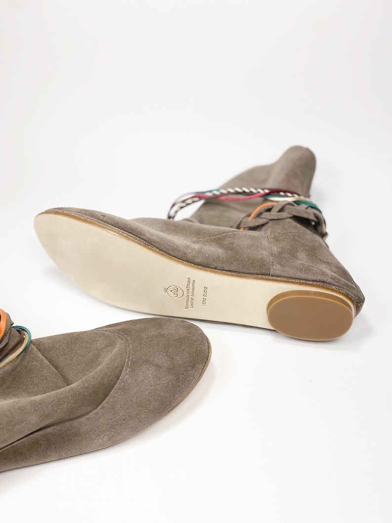 May include: A pair of gray suede ankle boots with colorful laces. The boots have a flat sole and a rounded toe. The text on the sole reads "Handmade and Design Luxury Accessories VFB 2020".