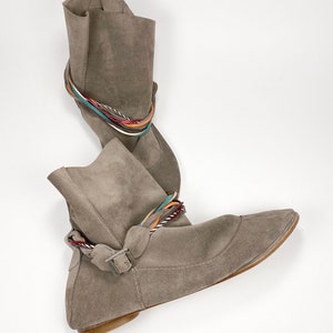 May include: A pair of gray suede ankle boots with a flat heel and a braided strap detail. The strap is made of multiple colors of cord.