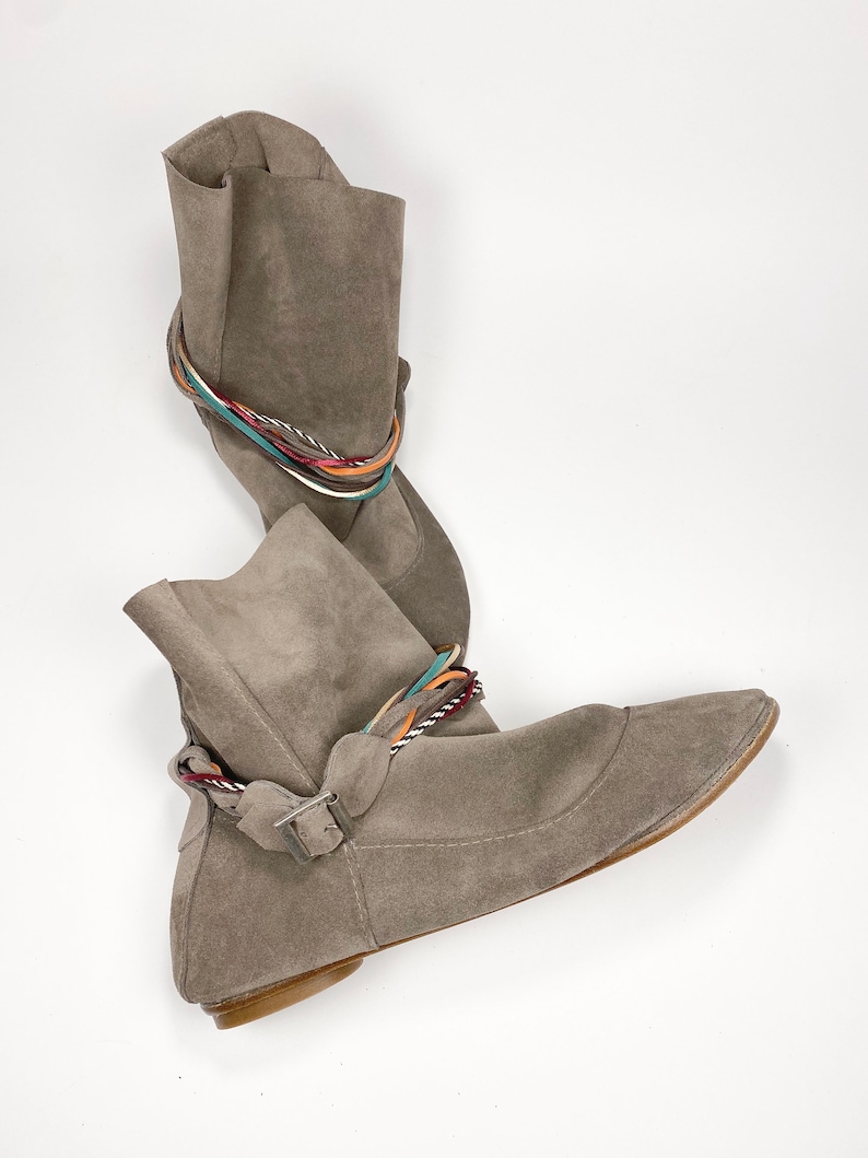 May include: A pair of gray suede ankle boots with a flat heel and a braided strap detail. The strap is made of multiple colors of cord.