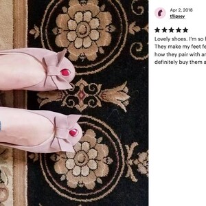May include: A pair of pink ballet flats with a bow on the toe. The shoes have a red accent on the bow. The shoes are on a patterned rug.