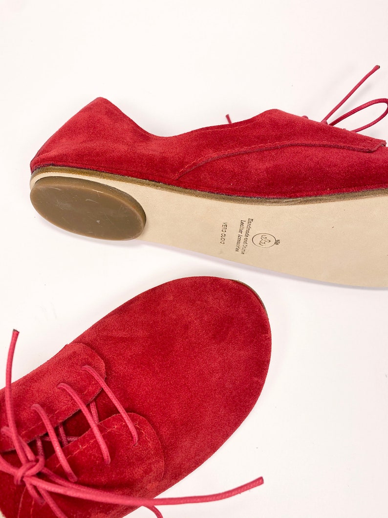 May include: Red suede lace-up shoes with a brown rubber sole. The shoes have a white leather label on the sole that reads "Limited edition Leather handmade 15/200".