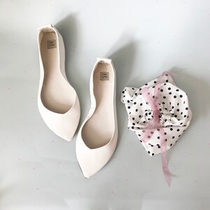 May include: Two pairs of white pointed-toe flats with a white fabric lining. The shoes are on a light blue background with white polka dots. A white fabric bag with black polka dots and a pink ribbon is in the background.