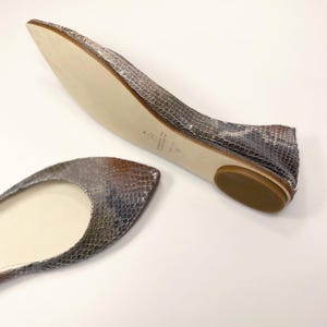 Peut inclure: Une paire de ballerines avec un motif peau de serpent dans les tons gris, marron et beige. Les chaussures ont un bout arrondi et un petit talon plat. La semelle est beige clair.