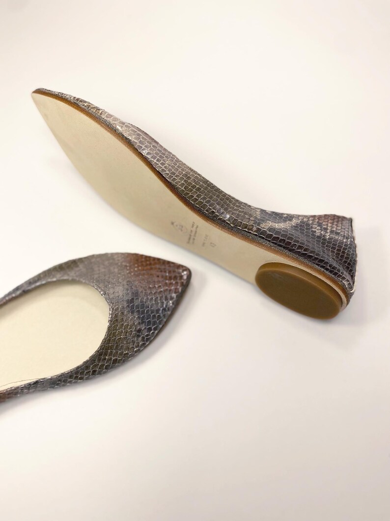 Peut inclure: Une paire de ballerines avec un motif peau de serpent dans les tons gris, marron et beige. Les chaussures ont un bout arrondi et un petit talon plat. La semelle est beige clair.
