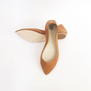 May include: A pair of brown leather ballet flats with pointed toes. The flats are laid out on a white surface.