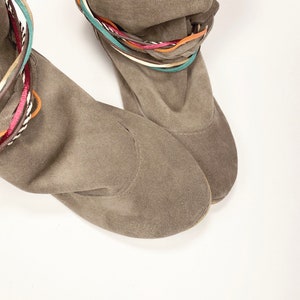 May include: A pair of brown suede ankle boots with colorful braided straps around the top of the boot.