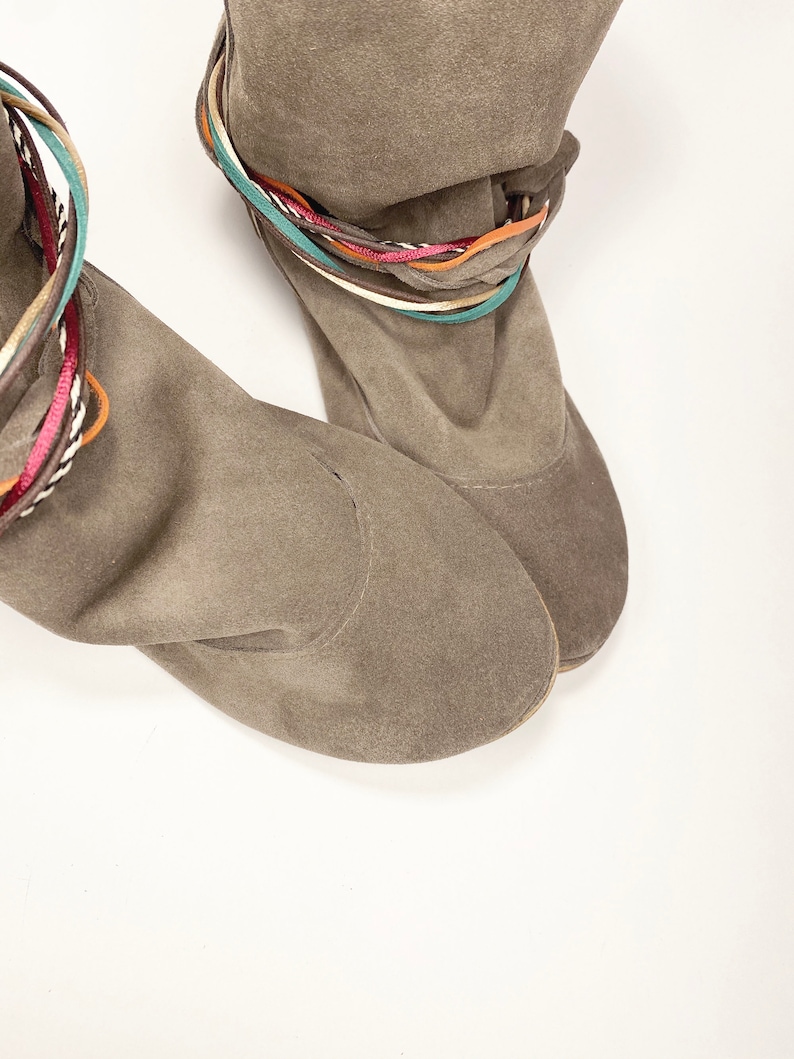 May include: A pair of brown suede ankle boots with colorful braided straps around the top of the boot.