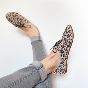 May include: A pair of leopard print lace-up shoes with a low heel. The shoes are worn by a person wearing grey jeans.