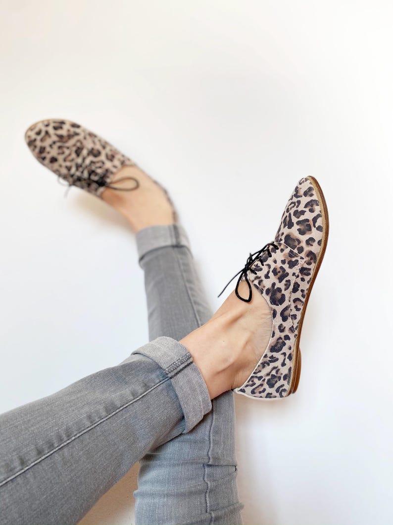 May include: A pair of leopard print lace-up shoes with a low heel. The shoes are worn by a person wearing grey jeans.