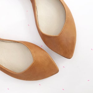 May include: A pair of brown leather ballet flats with pointed toes. The flats have a white leather lining.
