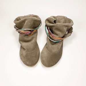 May include: A pair of brown suede ankle boots with colorful braided straps around the top of the boot.