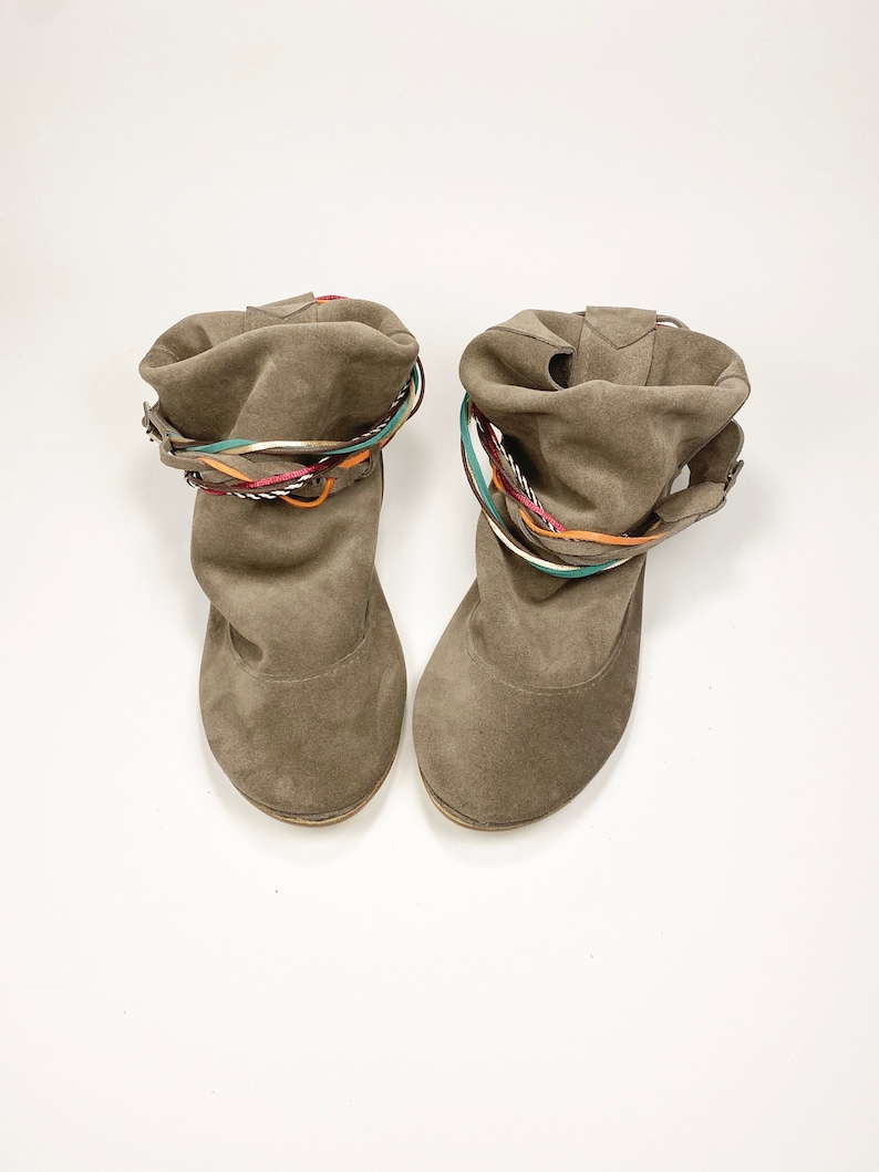May include: A pair of brown suede ankle boots with colorful braided straps around the top of the boot.
