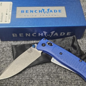May include: A blue Benchmade Bugout knife with a partially open blade, resting on a black pouch. The knife has a textured handle and a silver blade. A blue box with the Benchmade Knife Company logo is in the background.