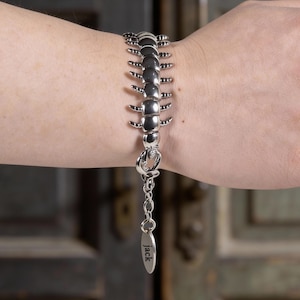 Puede incluir: Pulsera plateada con diseño de ciempiés. La pulsera presenta una serie de eslabones segmentados y redondeados con patas que sobresalen. Una pequeña etiqueta con el nombre "jack" cuelga del cierre.
