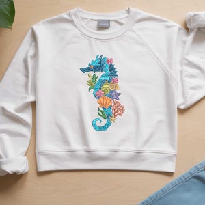 May include: White crew neck sweatshirt featuring a detailed seahorse design. The seahorse is blue with floral and seashell accents in pink, orange, and yellow. The design includes a starfish and coral. The sweatshirt has long sleeves.