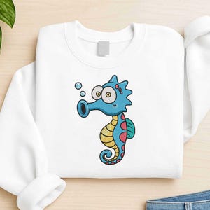 Puede incluir: Sudadera blanca con un diseño de caballito de mar de dibujos animados. El caballito de mar es azul con detalles amarillos y rosas, ojos grandes y burbujas. El diseño está centrado, creando un aspecto divertido y caprichoso.