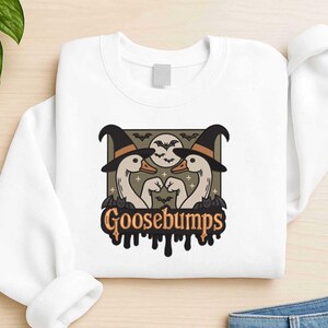 May include: White sweatshirt featuring a Halloween-themed graphic. The design includes two geese wearing witch hats, bats, a full moon, and the word "Goosebumps" in a dripping font. A fun and spooky design for the Halloween season.