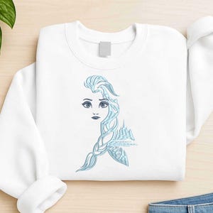 Puede incluir: Sudadera blanca con un diseño bordado en azul claro de la cara de una mujer, cabello suelto y un castillo. El diseño está en la parte delantera de la sudadera.
