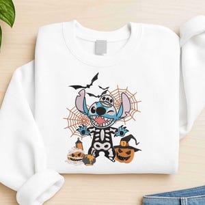 May include: White sweatshirt featuring a Halloween-themed graphic. The design includes a blue alien character in a skeleton costume, bats, spiderwebs, and jack-o'-lanterns. The sweatshirt is a crew neck style.